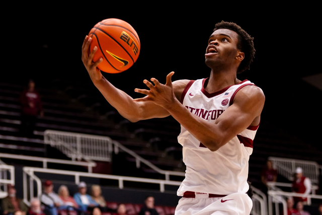 A basketball player wearing a Stanford jersey attempts a layup, holding the ball in one hand with eyes focused on the basket.